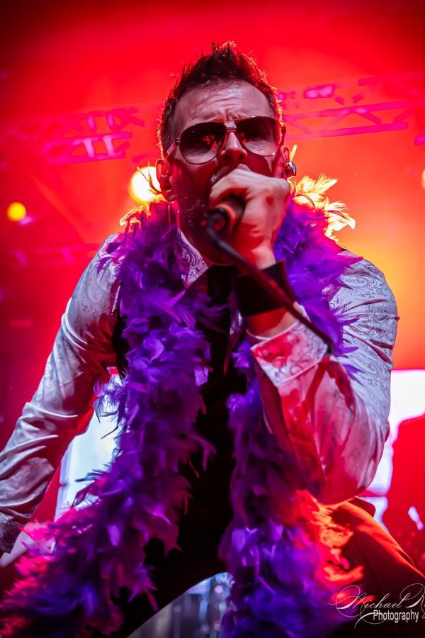 Singer in sunglasses and a purple boa, close on the mic under red light. Photo: Michael Key Photography.