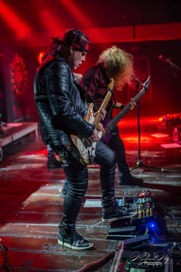 Two guitarists side by side at the front of a red-lit stage. Photo: Michael Key Photography.