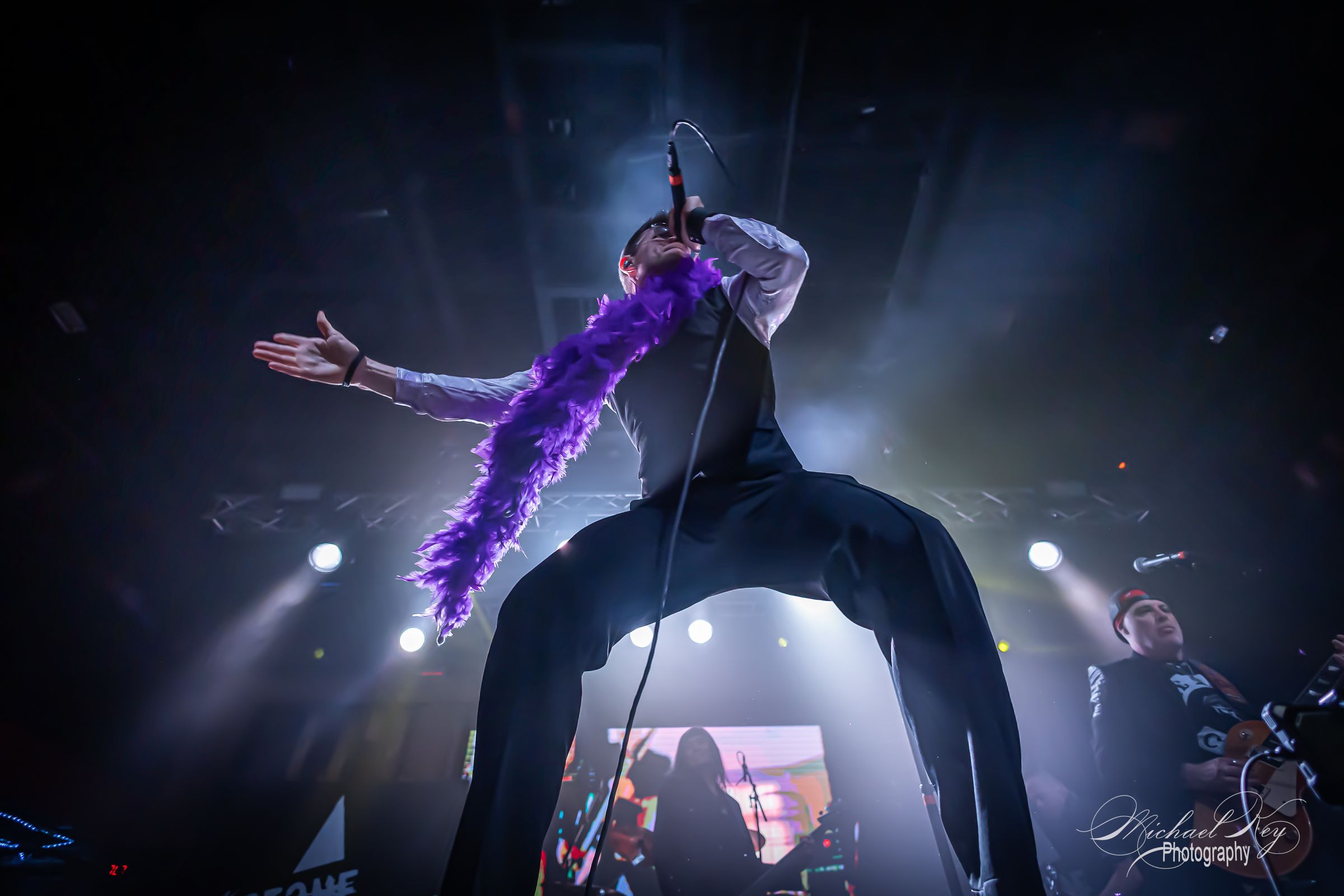 The singer backlit, arms up, a purple boa across his shoulders