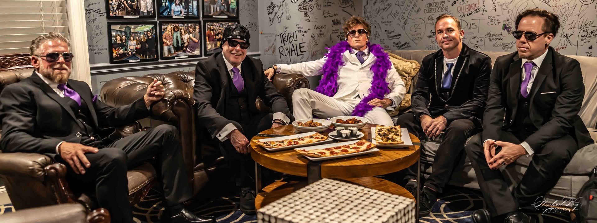 The five members of The Stone Temple Pilots Projekt backstage on leather couches, the singer in a white suit and purple boa, the others in black suits and purple ties
